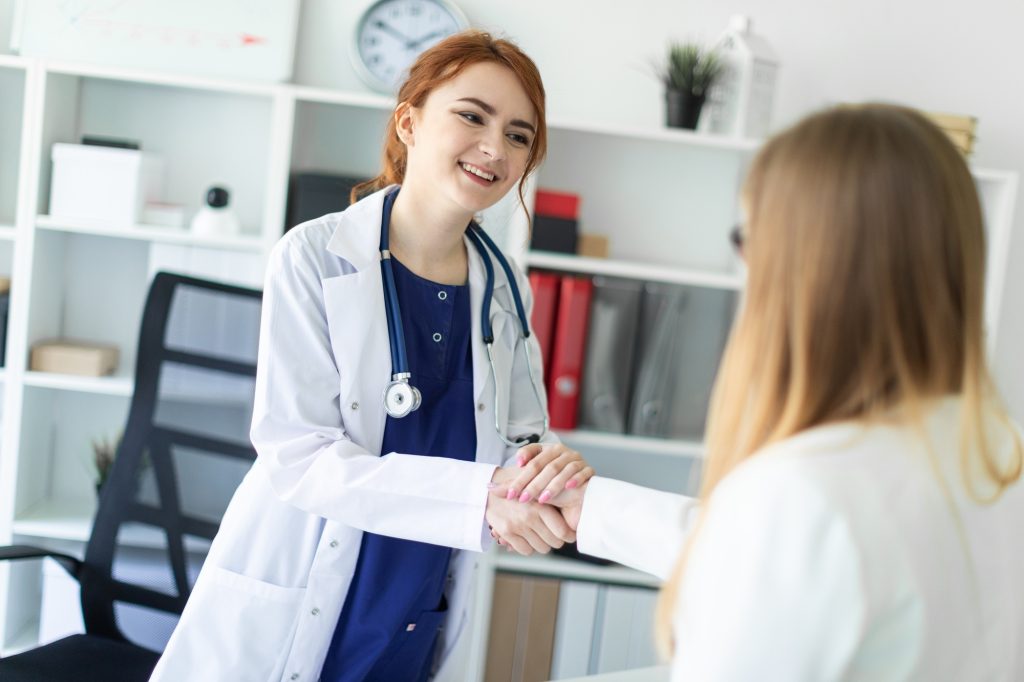 A beautiful young girl in a white coat is standing near the desk in the office and shaking hands